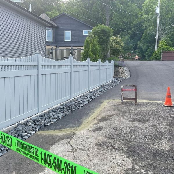 White fence along a driveway with rocks. A house is in the background. Construction cones are in place.