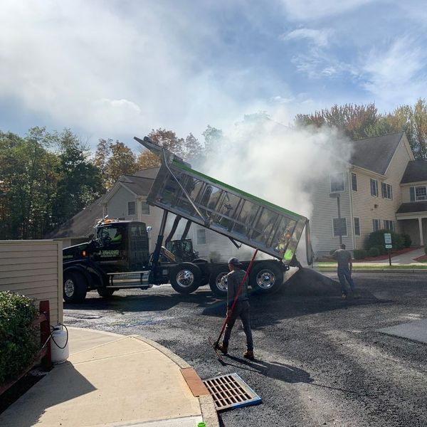 Asphalt being dumped from a truck onto a road by workers. Buildings in background.
