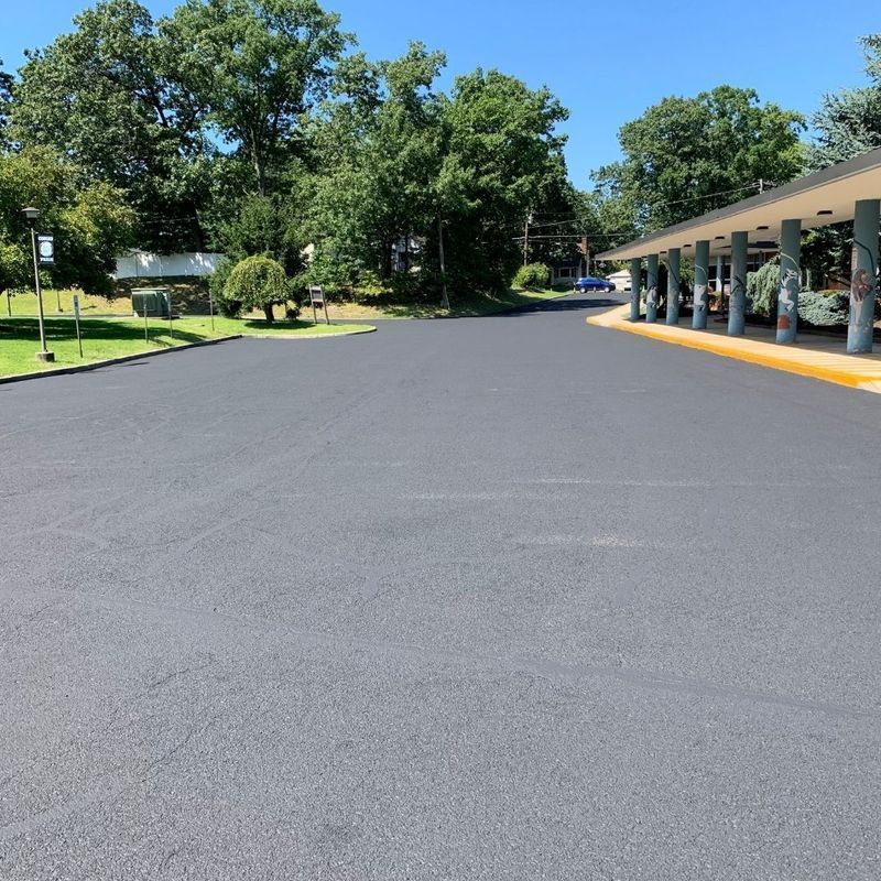 Newly paved asphalt parking lot with a covered area on the right and trees in the background.