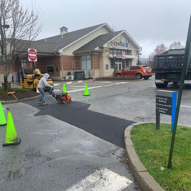 Man compacting asphalt in a bank driveway with cones and a truck, cloudy day.