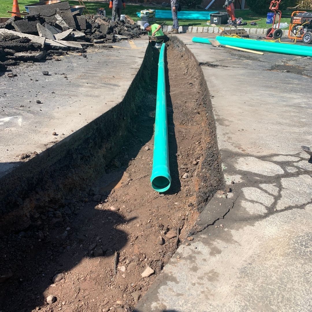 Trench dug in asphalt with a long green pipe laid in it; workers in background.