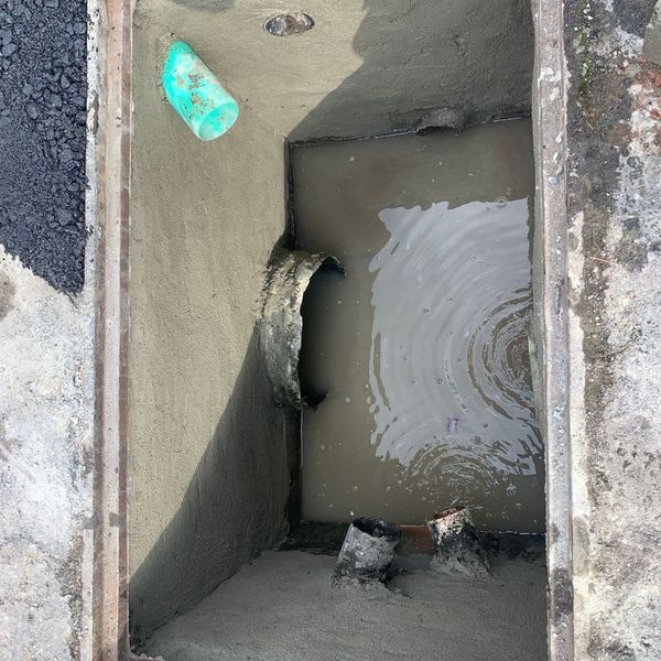 Open manhole with pipes and standing water, a plastic cone lies inside.