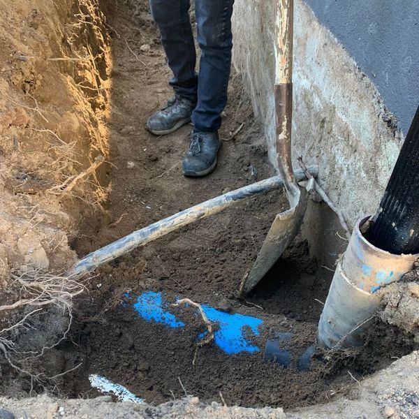 Person digging in a trench near a blue pipe, using a shovel. Soil, concrete, and black hose visible.