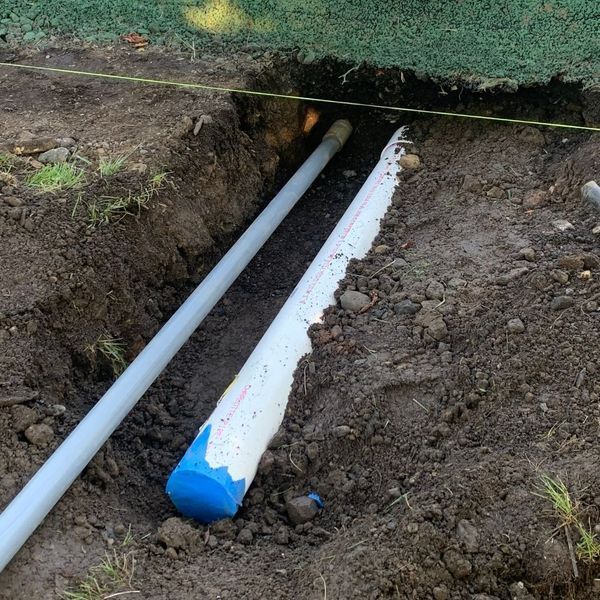 Pipes in a trench. One is white, and the other is light gray. The ground is dirt and grass.