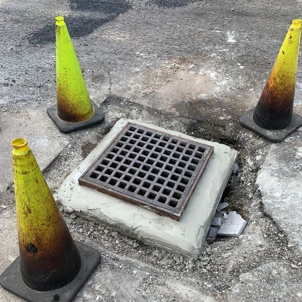 Open sewer grate on a paved surface, surrounded by three traffic cones.