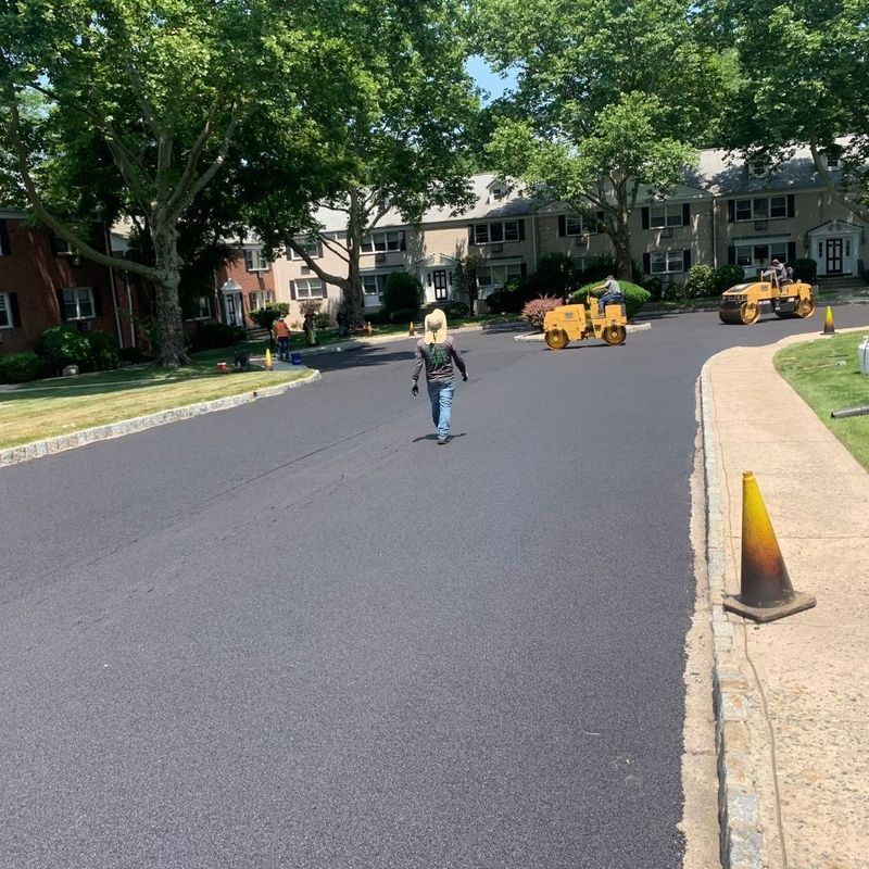 Street resurfacing; worker walks on freshly paved asphalt, with road rollers in background.