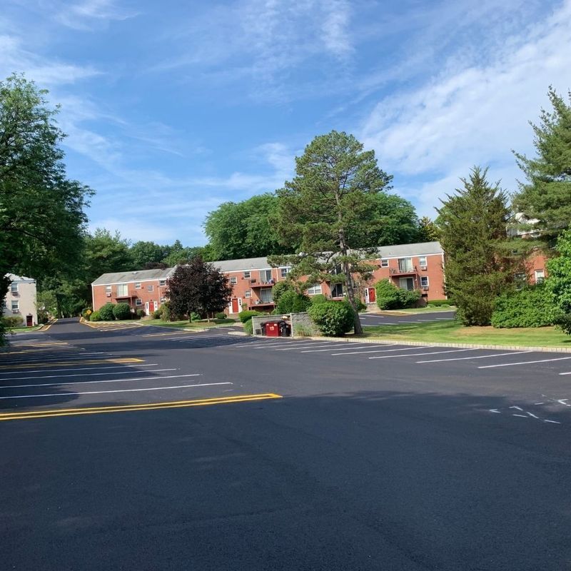 Apartment complex with red brick buildings, surrounded by trees and a parking lot on a sunny day.