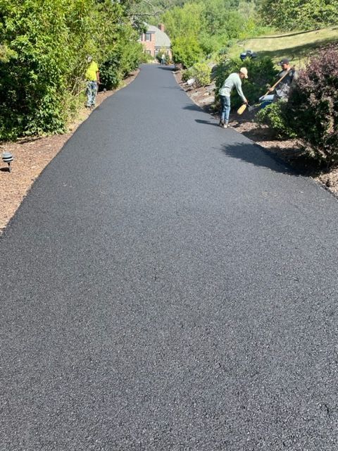 Workers paving a long, black asphalt driveway. One worker smooths edges with a broom.