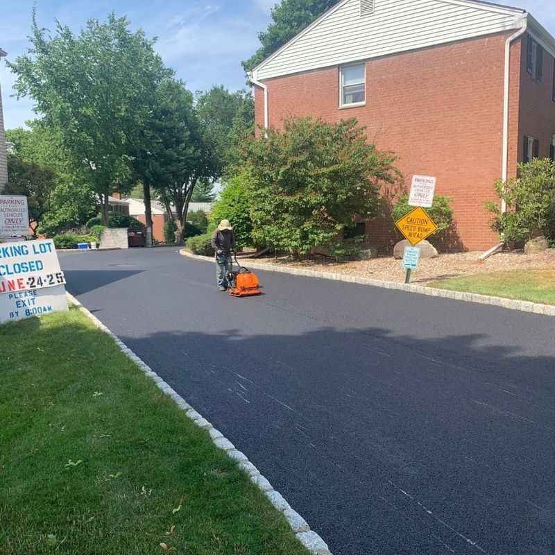 Worker compacting fresh asphalt on a parking lot; brick building, green grass, and trees in background.