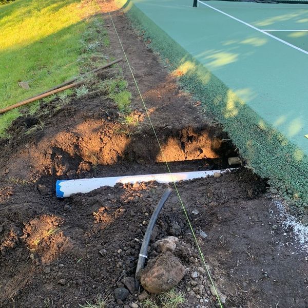 Trench dug beside a tennis court with buried pipe visible, yellow string guides, and green grass.
