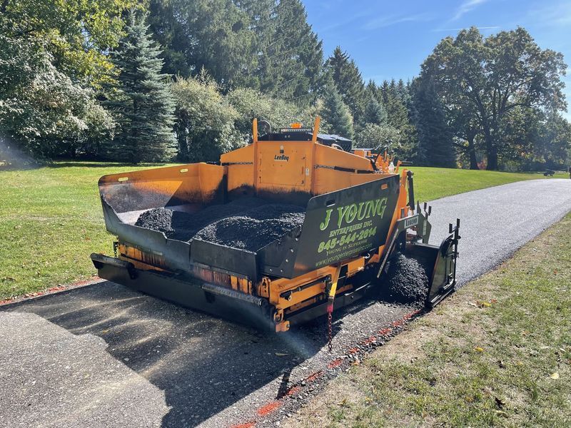 Asphalt paving machine laying blacktop on a road; setting is a grassy area with trees.