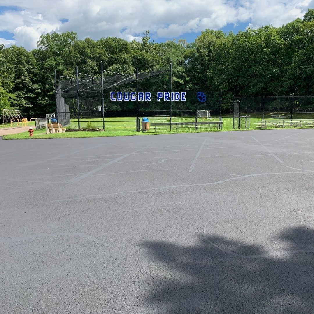 Black asphalt parking lot in front of a fenced baseball field, trees in the background, blue sign reads 