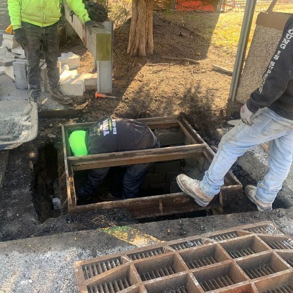 Workers in a pit; one inside, two outside. They are near a metal grate.