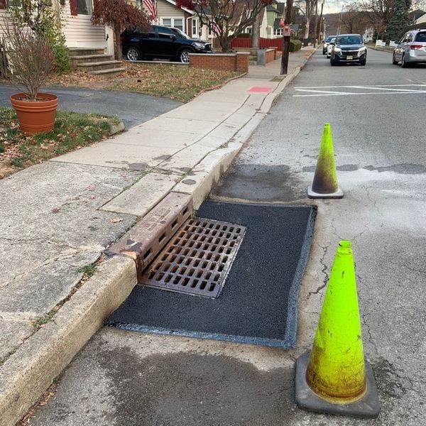 Street drain with metal grate, covered with asphalt. Two traffic cones warn of hazard.