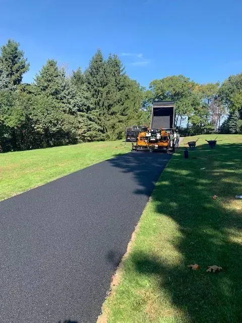 Asphalt driveway being paved; blacktop surface, green grass, trees, and construction vehicle.