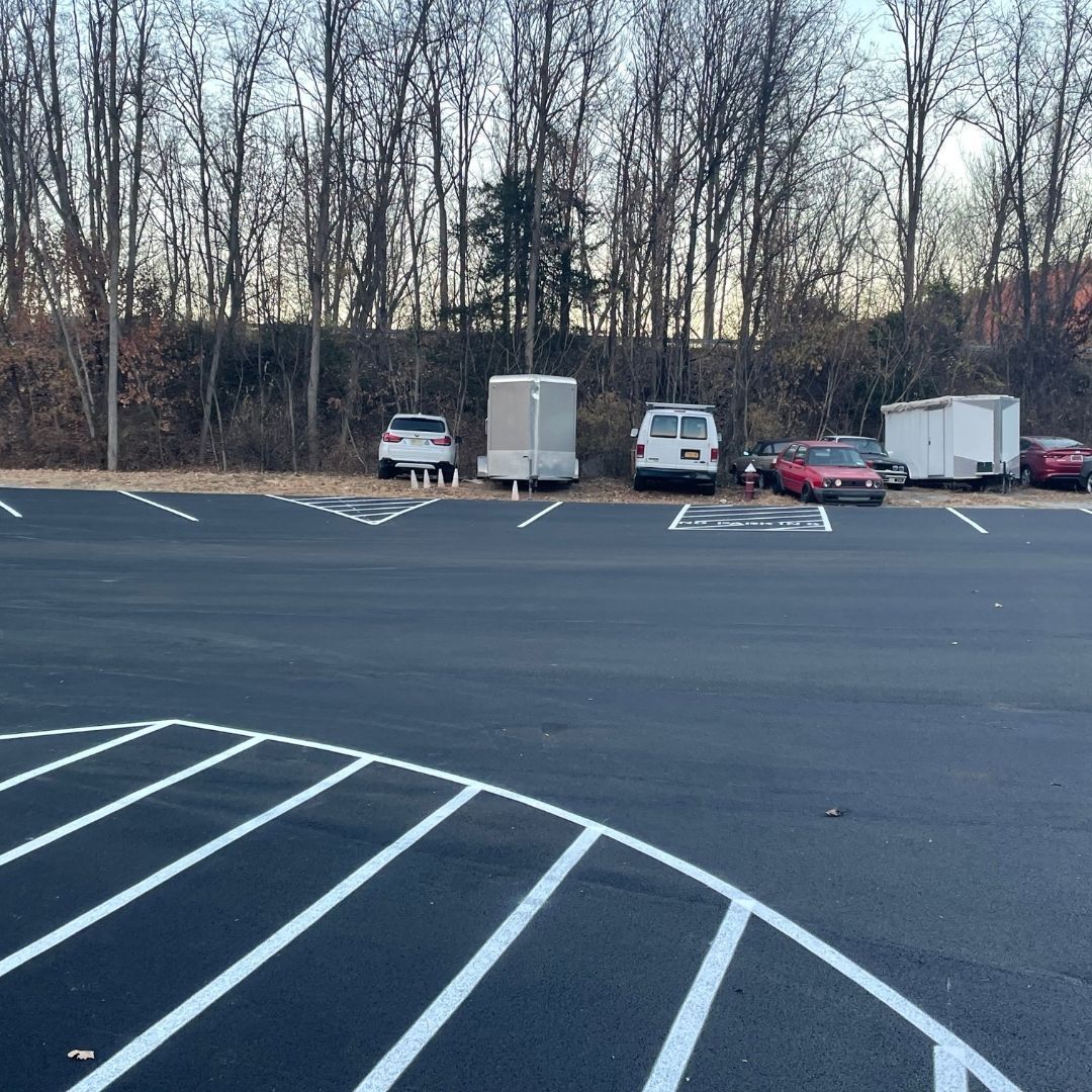 Paved parking lot with parked vehicles and trailers in front of a treeline.