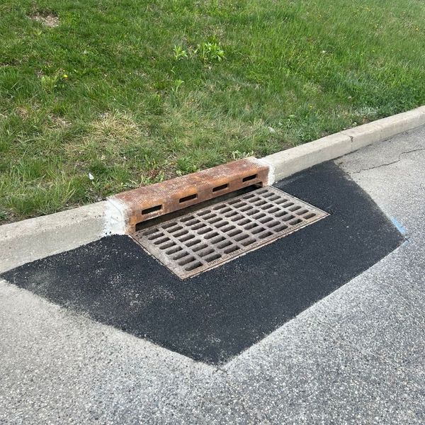 Drainage grate on asphalt by curb and grass. Metal grate, rectangular with square holes.