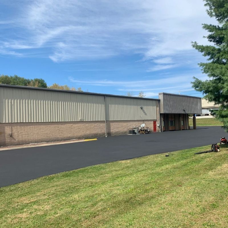 Building with corrugated metal siding and brick base, new asphalt, and green grass on a sunny day.