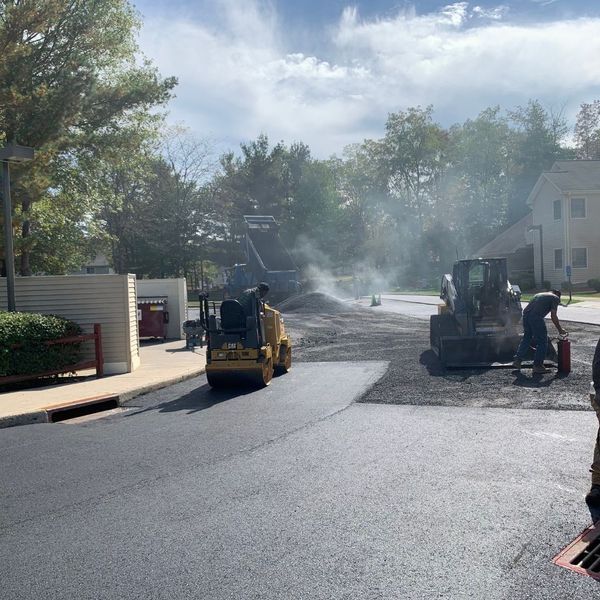 Asphalt paving in progress on a road; machinery includes a roller and a small bulldozer; a worker is present.