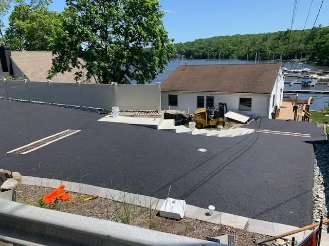 Newly paved asphalt parking area next to a waterfront home. A small construction vehicle sits on concrete steps.