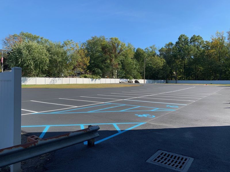 Empty asphalt parking lot with blue lines, bordered by a white wall and trees under a blue sky.
