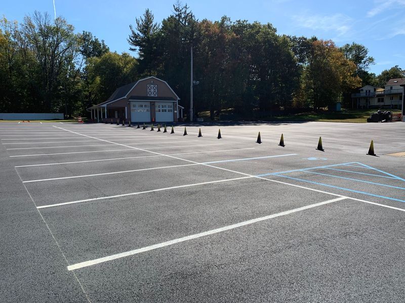 Empty asphalt parking lot with painted lines and orange cones. Building and trees in the background under blue sky.