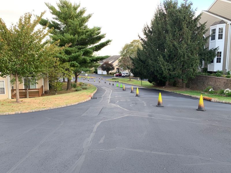 Asphalt road with orange cones, flanked by trees and buildings, possibly for road work.