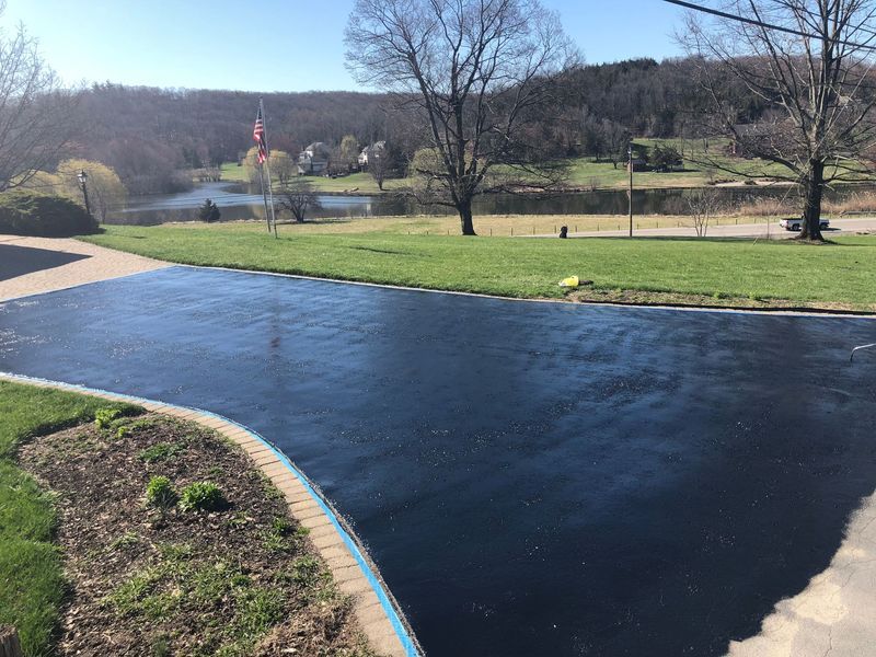 Black asphalt driveway next to green lawn, with a lake and trees in the background under a blue sky.