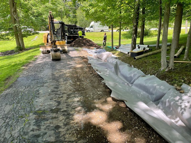 Excavator on a driveway digging up asphalt next to a white tarp and pipe rolls, trees in background.