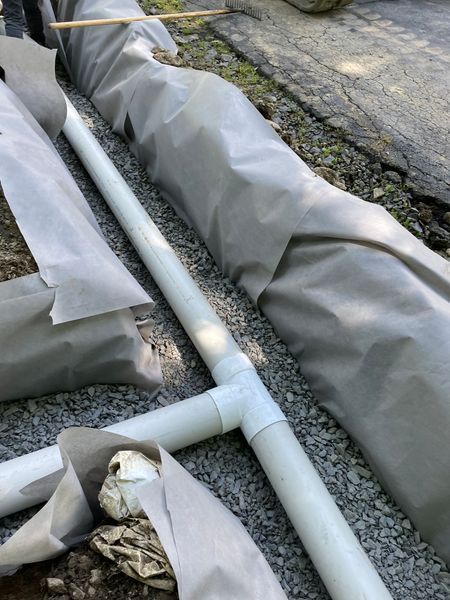 White PVC pipe and fittings installed in a gravel trench, wrapped in grey fabric.