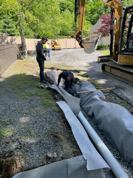 Drainage system construction: black fabric-lined trench with gravel and a white pipe along a roadside.