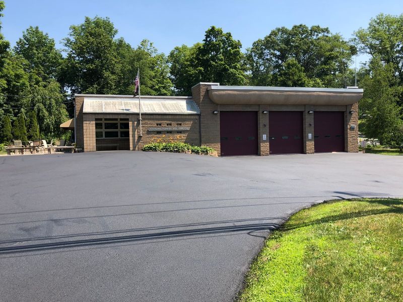 Fire station with three bays, brick exterior, dark red doors, flag, asphalt driveway, green foliage.