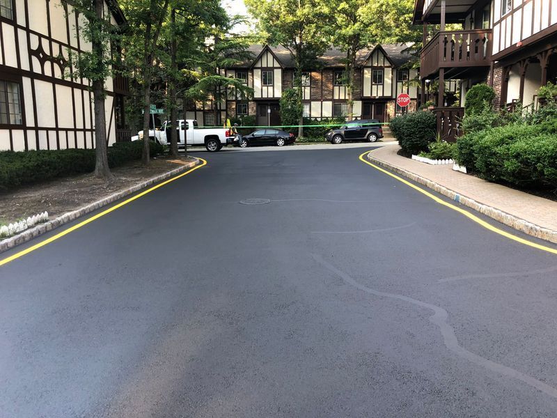 Newly paved dark asphalt road with yellow lines, in front of Tudor-style buildings and landscaping.