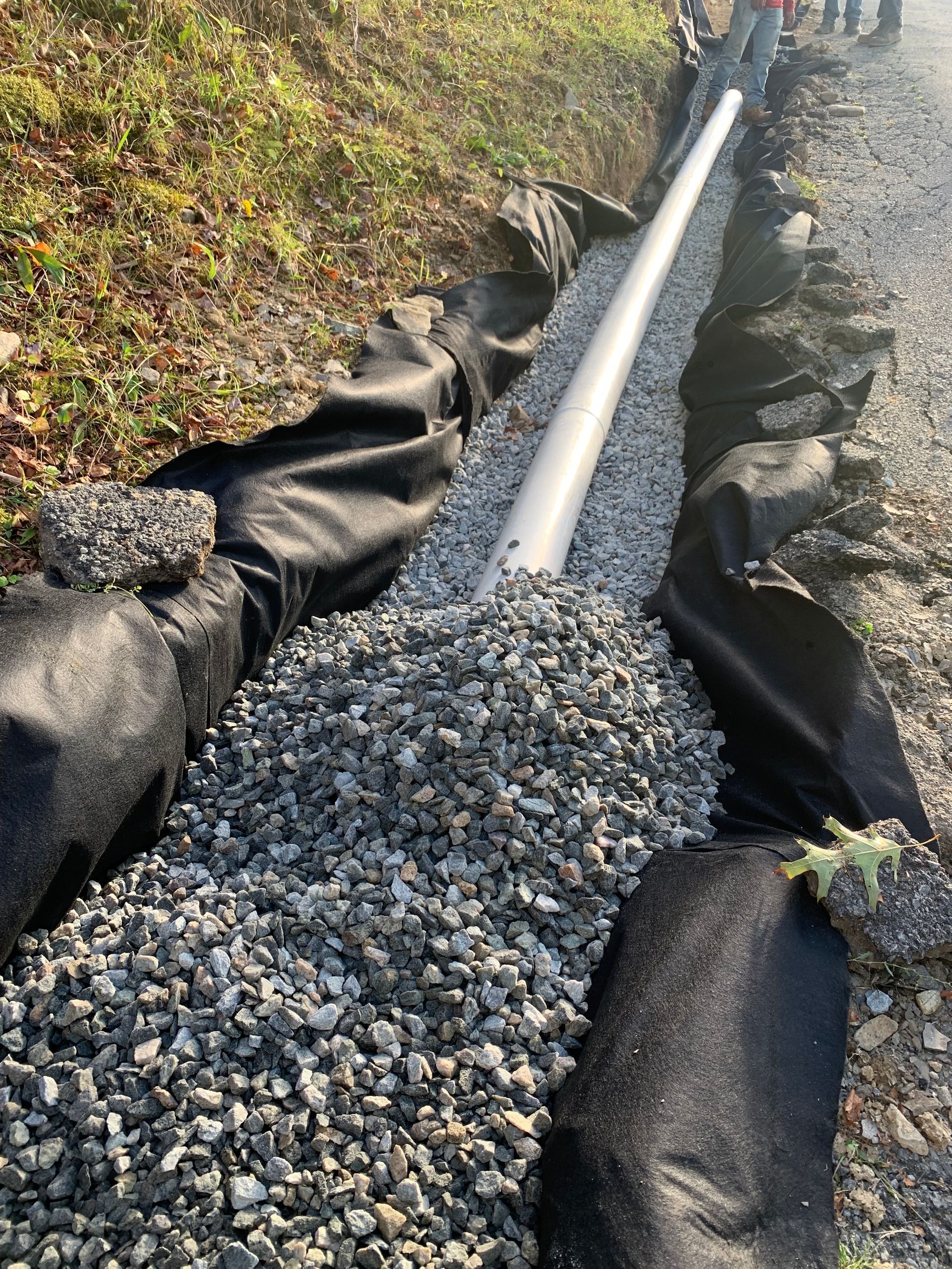 Trench with drainage pipe, gravel, and black fabric alongside a road.