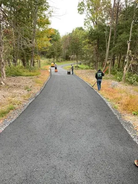 Newly paved asphalt driveway with workers; trees border the sides, overcast.