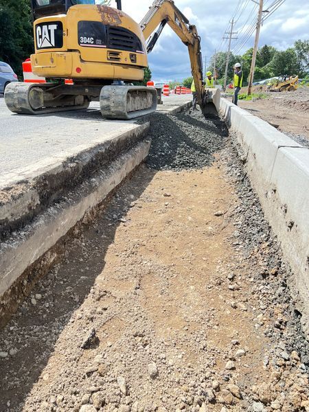 Excavator working on road construction, digging trench along curb; gravel and dirt visible; sunny day.