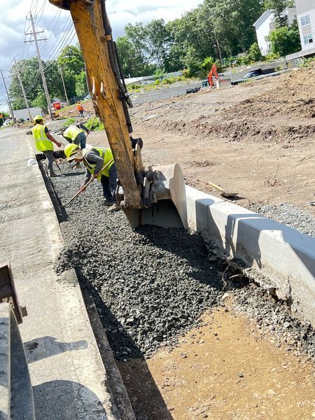 Construction workers filling a road gutter with gravel, using an excavator and hand tools, outdoors on a sunny day.