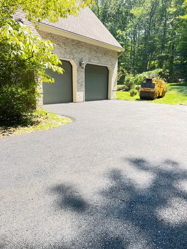 Asphalt driveway leading to a house with two gray garage doors. A yellow road roller sits on the grass.