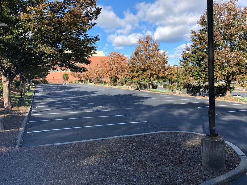 Empty asphalt parking lot with white lines, trees with brown leaves, blue sky, and a building in the background.