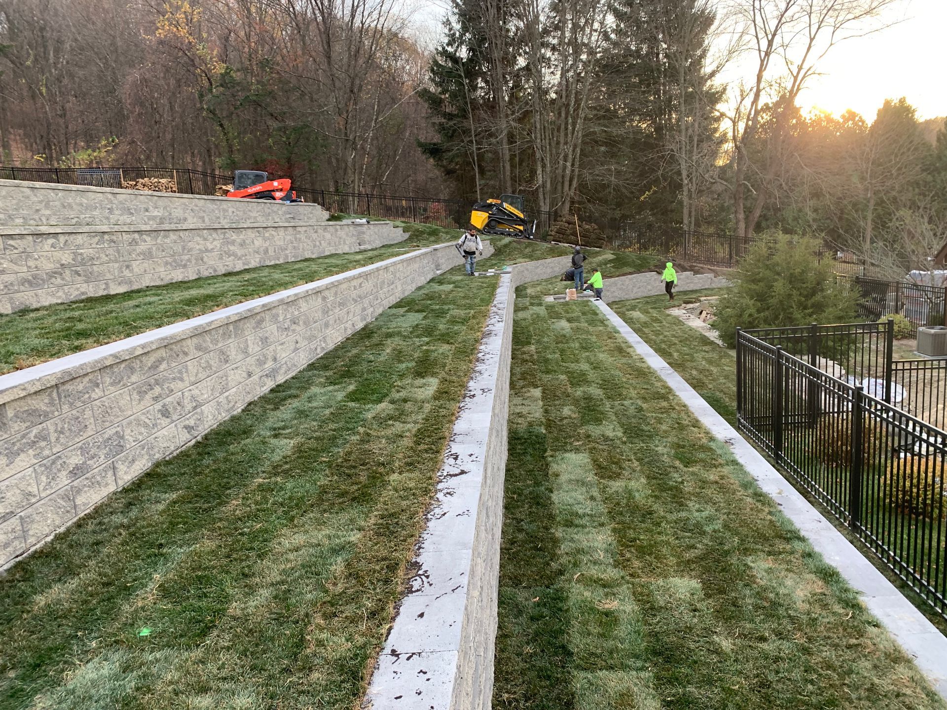 A tiered, newly landscaped hillside with retaining walls and sections of fresh turf, with workers visible in the distance.