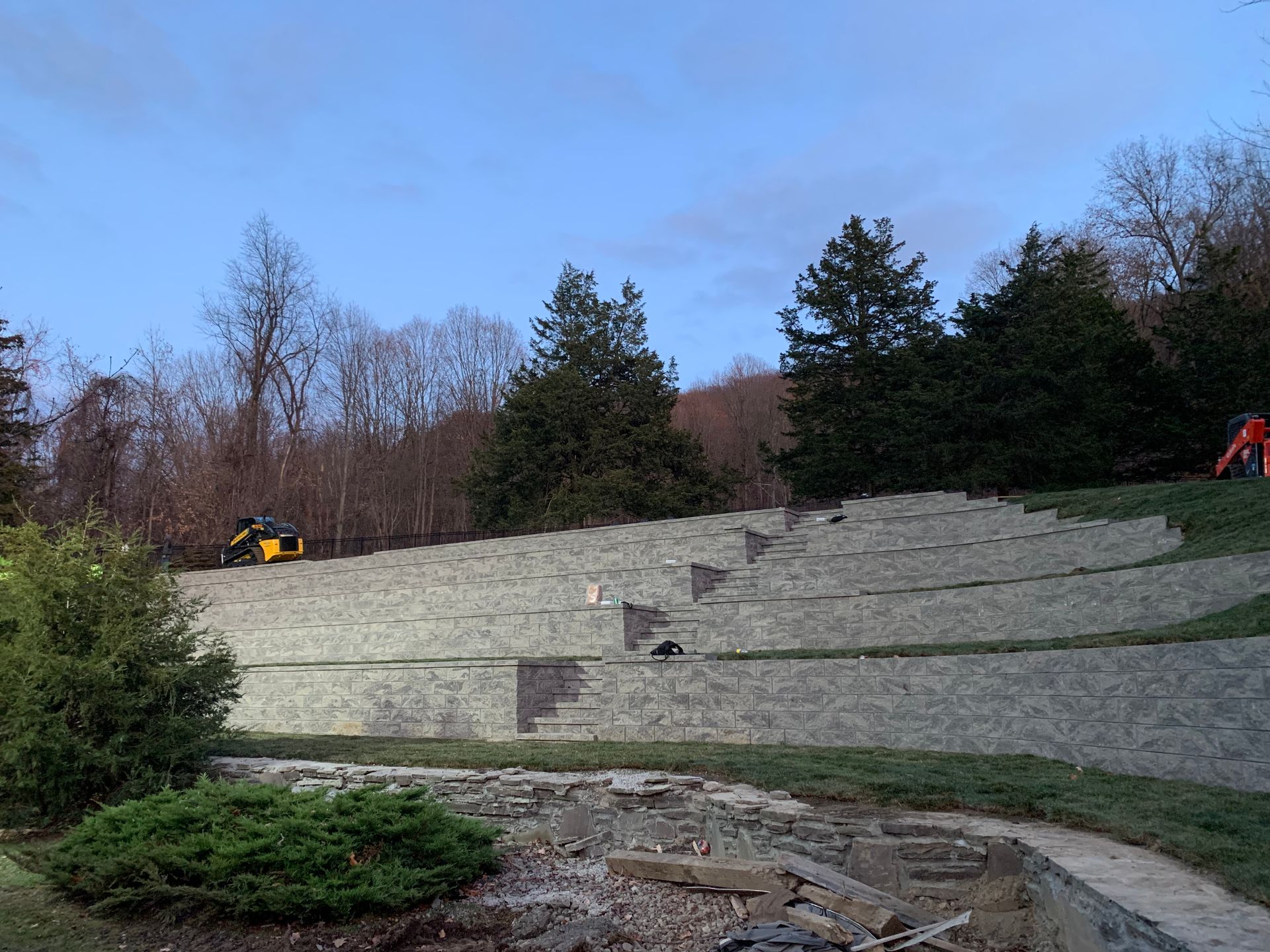 A multi-tiered stone retaining wall built into a grassy hill, with trees in the background and construction equipment nearby.