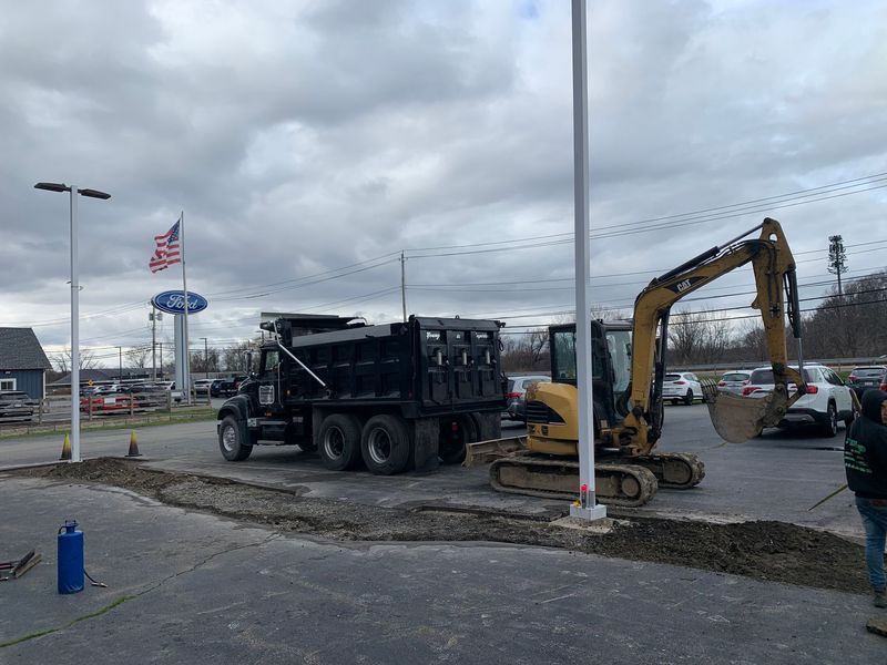 Excavator and dump truck at a Ford dealership; ground work is being done on a cloudy day.
