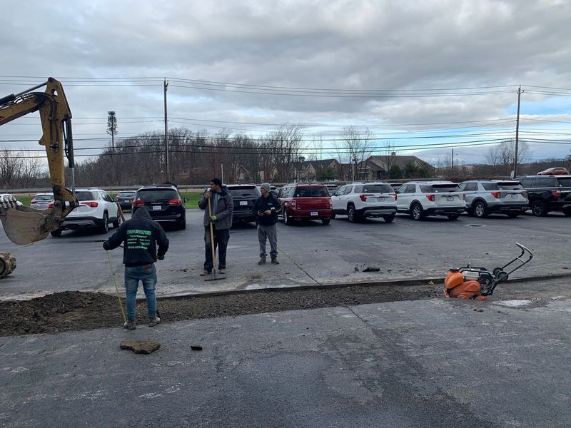 Construction workers repairing asphalt parking lot with equipment and parked cars visible.