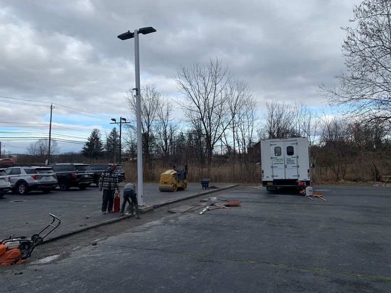 Construction site: workers near parking lot, tools, lights, truck, excavator, cloudy sky.