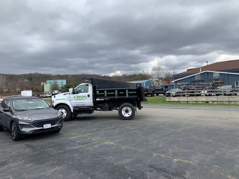 Dump truck parked in a lot, beside a gray SUV, under cloudy skies. Buildings and trees in the background.