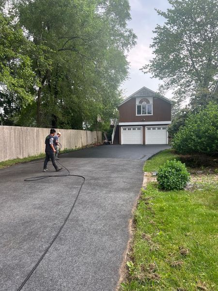 Man spraying sealant on a freshly paved driveway leading to a two-car garage. Trees and lawn are on either side.