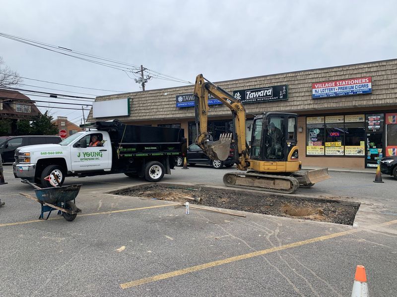 Construction in progress; excavator next to a dump truck at a strip mall. A large hole is in the pavement.