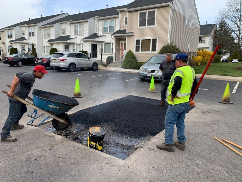 Workers patching asphalt in a parking lot. Two men use a wheelbarrow, one wears a safety vest. Buildings in background.