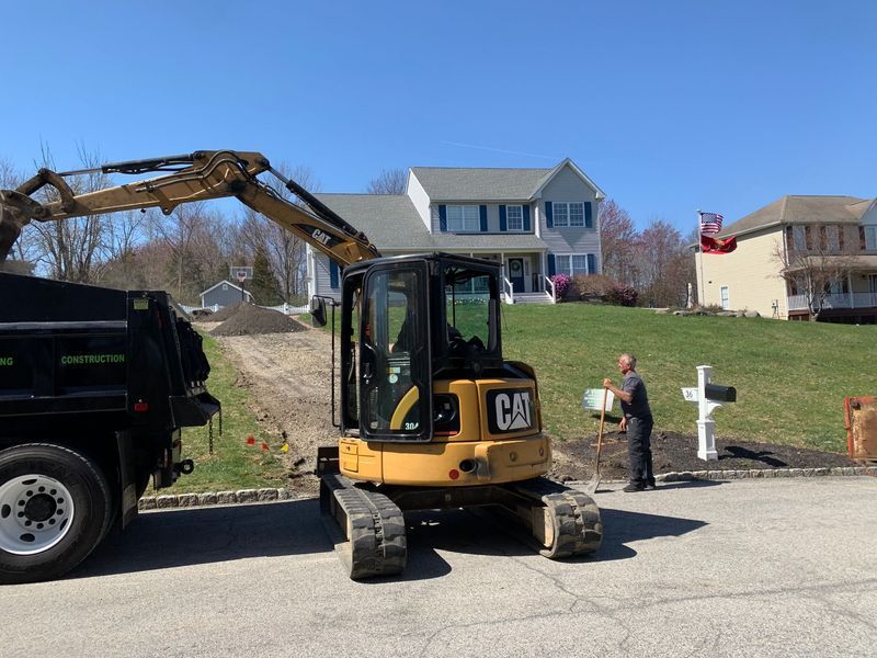 Yellow excavator loading a dump truck on a street in front of two houses. A man stands nearby.