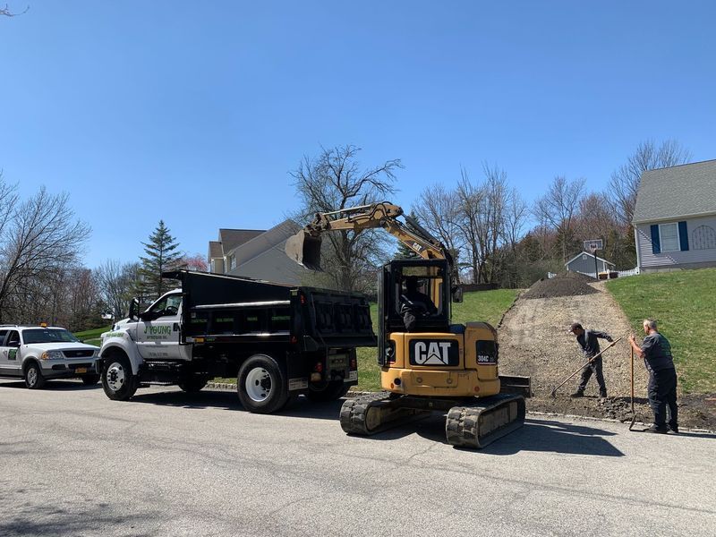 Excavator loading a dump truck on a driveway, two workers shoveling. Blue sky, suburban setting.